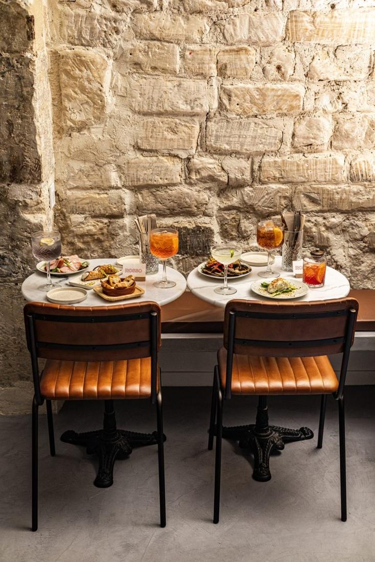 Tables en marbre, chaises en cuir cognac et spritz devant le mur de pierre